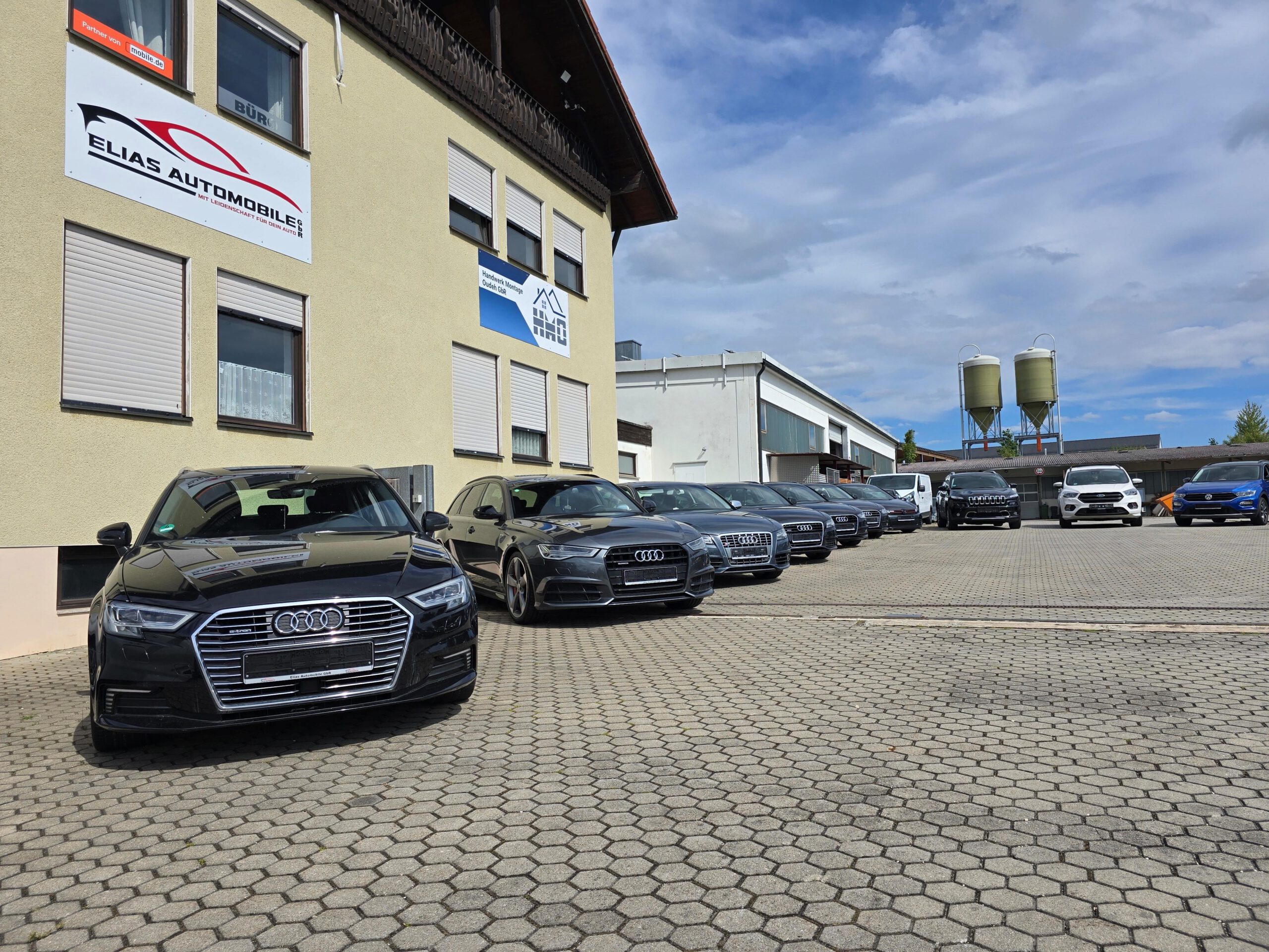 Mehrere schwarze Audi-Modelle stehen vor dem Autohaus aufgereiht, daneben weitere Fahrzeuge auf dem Hof.
